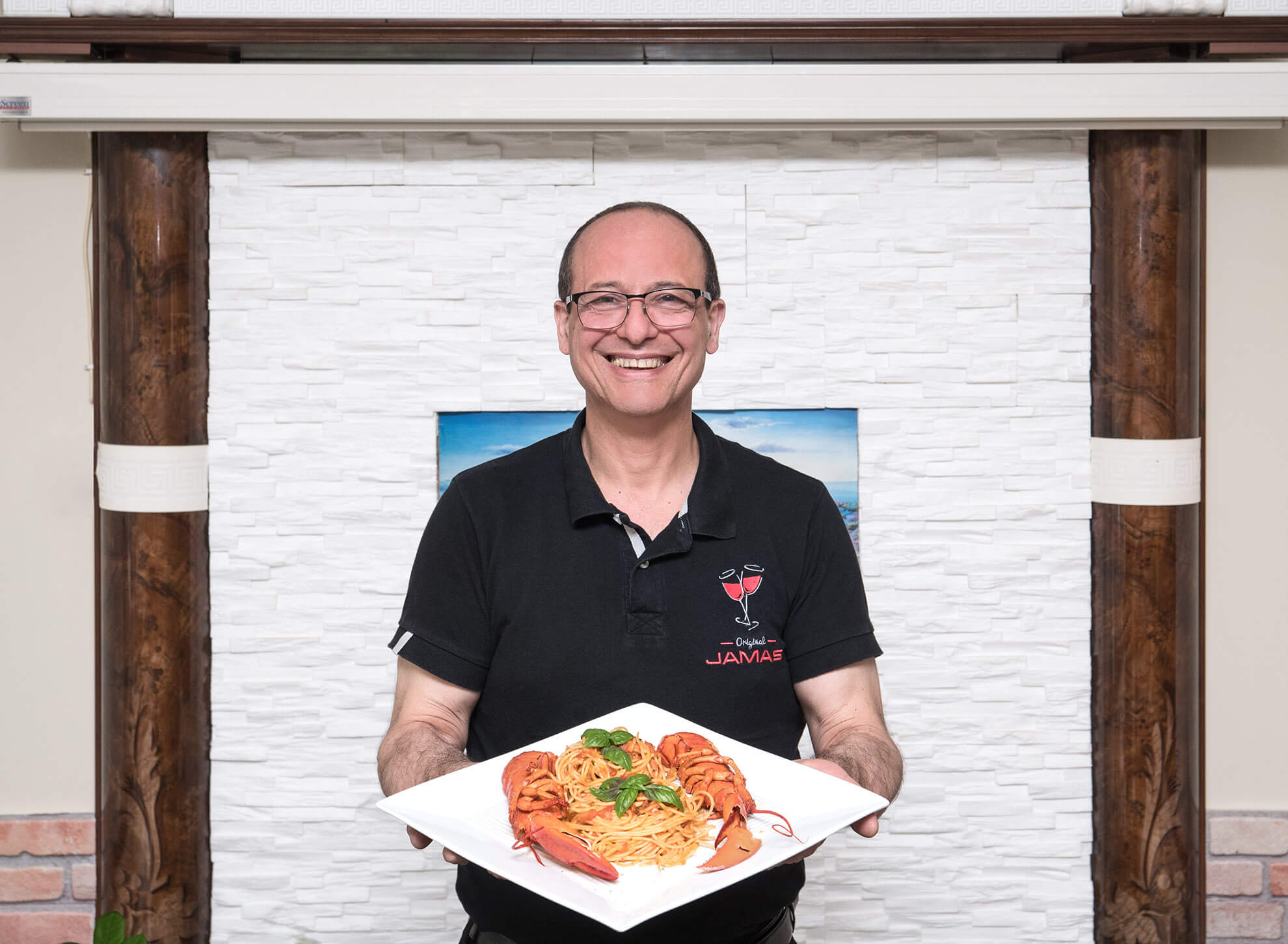 Der Besitzer des griechischen Restaurants Jamas präsentiert lächelnd ein griechisches Nudelgericht mit Meerestieren.