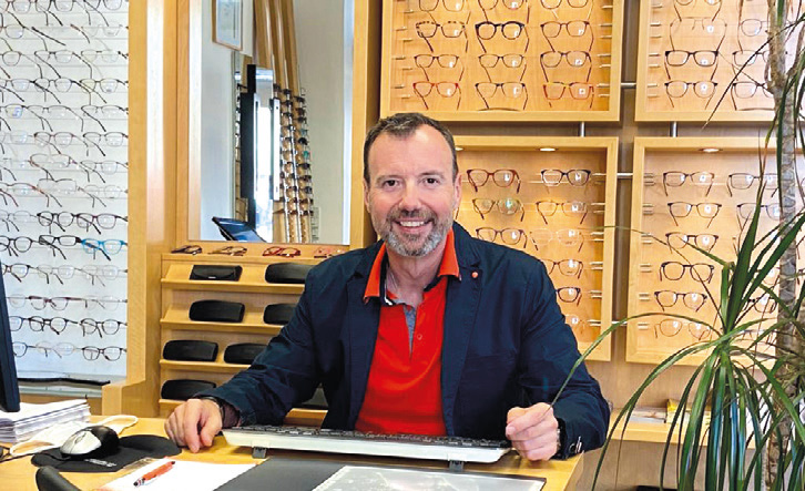 Optiker Walter Karall freundlich lächelnd an seinem Schreibtisch im Geschäftslokal. Im Hintergrund eine große Auswahl an Brillen.