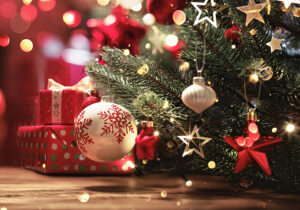 Im Vordergrund der untere Zweig eine Christbaumes mit einer silberenen Christbaumkugel. Dahinter einige rot verpackte Weihnachtsgeschenke.