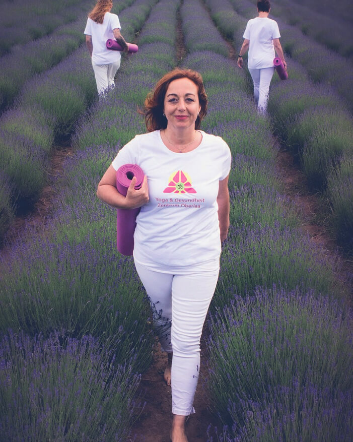 Yogalehrerin Nathalie Tisseyre geht in weißer Kleidung selbstsicher mit einer Yogamatte unter dem Arm durch ein Lavendelfeld.