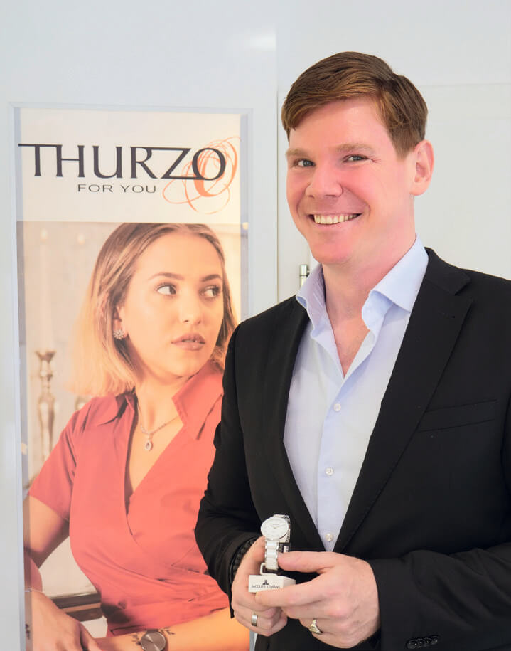 Philipp Thurzo lächelnd vor einem Plakat für Thurzo-Juwelen.